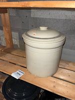 Full view of white crock with lid on wooden shelf, label showing lot number on shelf.