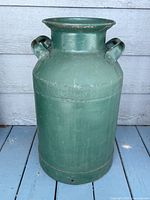 Front view of the tall green painted metal milk can with handles on each side, standing on a painted wood floor.