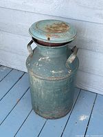 Full view of vintage metal milk can standing on a painted blue wooden floor against a light-colored wall, showing two large handles and attached lid slightly tilted.