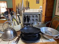 Wide view showing stainless steel kettle, metal colander, black knife block with knives, baking tray and rack, wooden handled sharpening steel rod, and white utensil caddy with black and metal kitchen utensils.