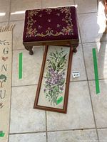 Small footstool with maroon needlepoint topped surface and floral design next to a framed floral needlepoint textile artwork, both on floor tiles. The footstool has wooden legs and the framed artwork is long and narrow.