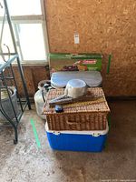 Lot contents showing stacked items including cooler, wicker basket, tin water ladle, propane canisters, and boxed Coleman stove behind.