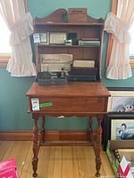 Full view of the wooden small desk with hutch placed against a wall, showing open shelves with various objects on the hutch, and the desk with turned legs under it.