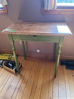 Small wooden table with green painted legs and natural wood top shown from front signal angle, highlighting wear and surface condition.
