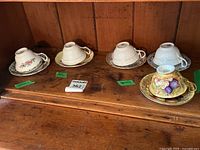 All five teacups and saucers displayed upside down on wooden shelf with brand labels visible.