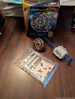 Front view of toy beyblade, launcher, instruction sheet in plastic bag, and original box on table.