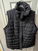Front view of black quilted vest showing high collar, zipper closure, and sleeveless design.