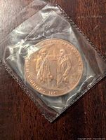 Bronze commemorative coin in plastic sleeve showing raised images of John A. Macdonald and George Cartier with text and date 1847.