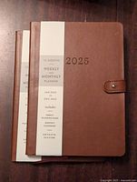 Photo showing two brown leather planners for 2025 with snap closure and labels indicating weekly and monthly planning pages.