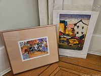 Both artworks side by side leaning against wall, one framed and one unframed.