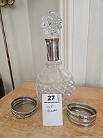 Glass decanter with sterling silver collar and stopper, diamond cut pattern, displayed with two silver coasters on either side and white background