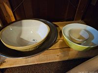 Set of four mixing bowls including two white pottery bowls with yellow rims, one yellow Pyrex bowl, and one large metal bowl stacked on a wooden shelf.
