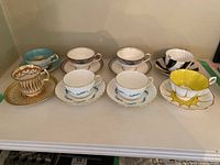 Photo showing 7 vintage china cups and saucers arranged in a row, showcasing various colors and design patterns including gold accents, turquoise, and yellow petal design.