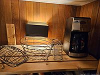 Photo showing metal kitchen accessories including a fish-shaped rack, a fruit basket, cutlery holder, and the coffee maker on a wooden shelf.