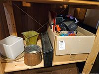 Photo of wooden shelf holding a copper pot, black metal mailbox, two plant pots (white and green), and a cardboard box with assorted small items