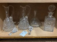 Photo of 4 cut glass decanters on wooden shelf, 3 lids visible, clear intricate cut designs.