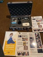 Opened metal case containing camera components for dental photography, foam insert with lenses, camera, USB cable, and memory card reader. Instruction booklets and manual laid flat next to case.