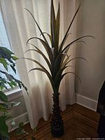 Full view of the faux tropical plant showing the textured trunk and long, pointed leaves against a curtain background.