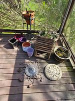 Collection of ceramic plant pots in various sizes and colors arranged on wooden deck and plant stands, including pink, purple, terracotta pots and metal sun plaques on deck.
