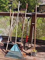 Full view of seven garden tools: two rakes, shovel, pitchfork, hand cultivator, spiked aerator, and hoe standing against railing outdoors.