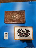 Photo showing both keepsake boxes on a blue surface: one Indigenous Raven design box signed by Clarence A. Wells, the other a large carved wooden box with mother-of-pearl inlay floral design.
