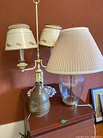 View of three lamps on a wooden surface against a reddish wall: a double-arm lamp with decorative beige shades, a green ceramic lamp base without shade, and a large clear glass lamp with a white pleated shade.