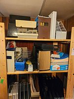Three shelves packed with various office supplies including file holders, paper trays, cardboard organizers, and an analog scale on the top shelf