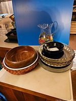 Photo of assembled lot showing glass pitcher, wooden bowls, various pie plates, small black bowls, clear glass platter, and woven base plate on countertop in front of blue background.