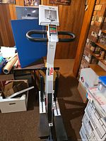 Precor exercise climber showing the full device with black handlebars and stepping platforms in a wooden-paneled room.