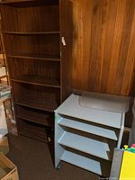 Large dark brown particle board shelving unit next to white metal shelving unit on casters, set against wood paneled basement wall