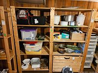 Close view of two connected wooden shelving units in a basement, holding containers, kitchenware, and boxes. The shelving is made of raw wood with visible screws and bolts for assembly.