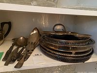 Photo showing several tarnished silver trays stacked with one lid having a handle and some silver serving utensils including salad servers
