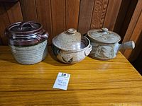 Three stoneware lidded casseroles on wooden surface against wood panel background. The casseroles vary in design and color with one large brown glazed piece, one speckled brown and beige piece, and one smaller patterned piece with a handle.