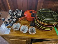 Overview of collection including Sunburst Canada fondue plates stacked, orange pot with wooden handle, black metal stand, Greek ceramic tea set, and three ceramic bowls.