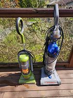 Two upright vacuum cleaners placed side by side outdoors on a wooden deck, showcasing the full view of both units including handles, bases, dust containers, and power cords.