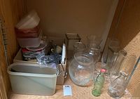 Photo of shelves with glass vases, glass pitcher, small green glass bottle, plastic containers and aluminum trays stacked, metal skewers and small kitchen accessories.