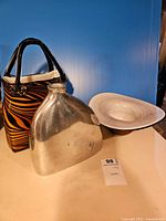 Photo showing three vases on a surface with a blue background: a shiny metal vase with a smooth surface and an irregular shape, a tall glass vase with black and orange tiger stripe and black handles, and a wide white and gray marble-glass vase with a broad lip.