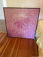 Framed square abstract artwork with textured pink pattern leaning against a wall on hardwood floor beside a bed.
