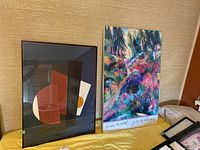 Overview showing two framed art pieces on a yellow surface against beige wallpaper: abstract geometric print and colorful nature-themed 'earth garden' print.