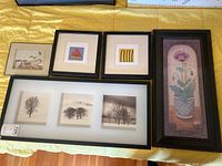 Five framed wall art pieces laid out on a yellow cloth, including a tall narrow floral painting, three small square original artworks (watermelon, asparagus, rustic wood), and a wide frame with three tree silhouette prints.