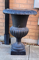 Front view of large black cast iron urn showing classical fluted design and scalloped rim, placed outdoors against brick wall.