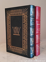 Two leather-bound books standing upright. The dark green book is 'Leaves of Grass' by Walt Whitman, and the red book is 'Kenilworth' by Sir Walter Scott. Both have gold decoration and gilded edges.