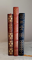 Three leather bound books standing upright showing spines with gold detailing and titles.