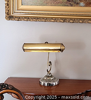 Front view of the brass piano lamp showing the elongated cylindrical shade with decorative ends, mounted on a curved arm attached to a silver-colored round base on wooden furniture beneath a framed picture.