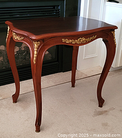 Side angle of a wooden accent table showing curved legs with gold ormolu mounts and a shaped top surface.