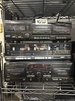 Three double cassette decks stacked on a metal shelf, showing front panels and control buttons.