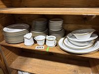 Full view of china set stacked on wooden shelf showing plates, bowls, teacups, creamer, sugar bowl and serving dishes with gold trim.