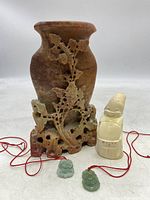 Full view of the soapstone vase with floral carvings, two jade pendants on red cords, and Billiken figurine side by side.
