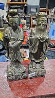 Photo showing two concrete Buddha-type statues with gold patina finish on a wooden surface surrounded by various items.