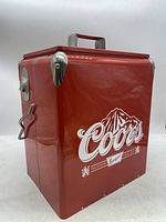 Red rectangular metal Coors Banquet cooler chest with white branding and mountain logo. Metal handle on top and metal corner protectors visible.